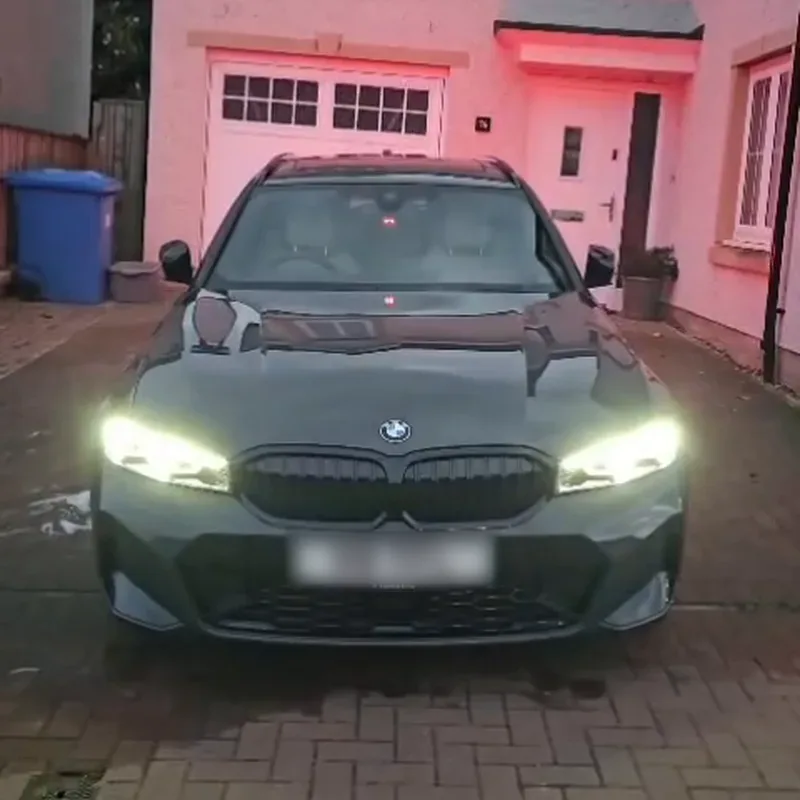 A grey BMW G20 320i on a driveway at dusk, its headlights on